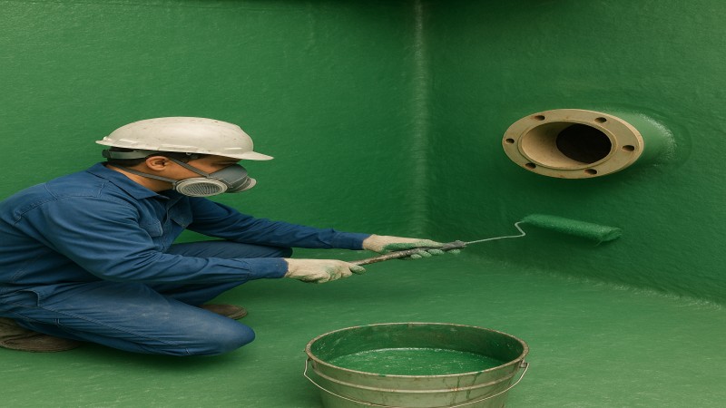 A worker in safety gear applies green GRP (Glass Reinforced Plastic) lining inside an industrial tank using a roller, with a bucket of resin nearby and a pipe flange visible on the wall.